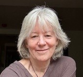 An older woman with grey hair wearing a grey blouse smiling at the camera